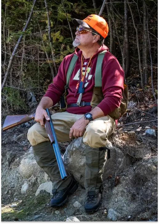 Man sitting on a rock in a forest, wearing an orange hunting cap and vest, holding a shotgun, representing outdoor hunting activities at RiverVail Ranch.