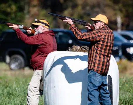 Two men aiming shotguns while upland bird hunting, surrounded by green grass and trees, with vehicles in the background, showcasing outdoor recreation at RiverVail Ranch.