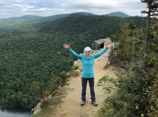Person celebrating at the edge of a scenic mountain overlook, surrounded by lush greenery and distant hills, embodying the spirit of outdoor wellness at RiverVail Ranch.