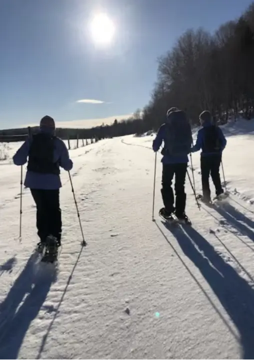 Three individuals snowshoeing on a snowy trail under a bright sun, surrounded by trees, showcasing outdoor wellness activities at RiverVail Ranch.
