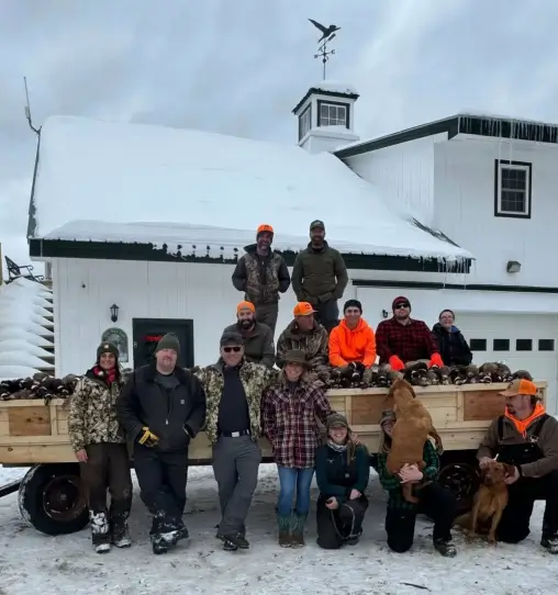 Group of hunters in winter attire posing on a snow-covered wagon filled with upland birds, with a rustic ranch building in the background, showcasing outdoor recreation at RiverVail Ranch.