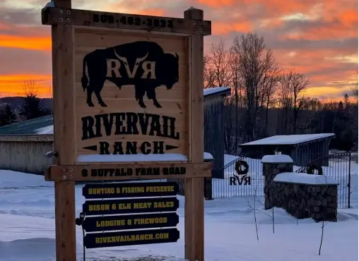 Sign for RiverVail Ranch with a bison silhouette, displaying information about hunting, fishing, and lodging, set against a winter sunset backdrop.