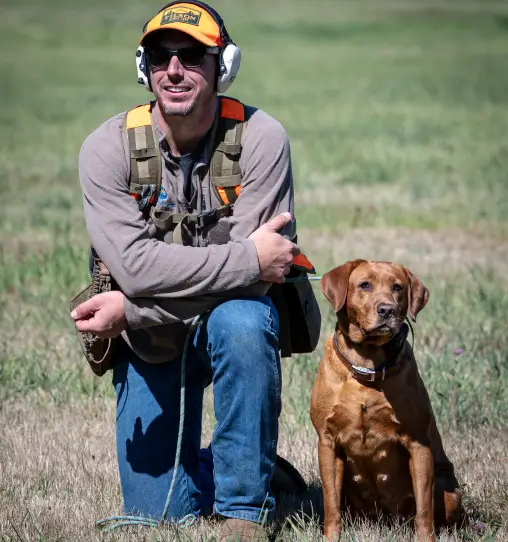 Man kneeling beside a dog in a grassy field, wearing hunting gear and ear protection, embodying outdoor activities at RiverVail Ranch.