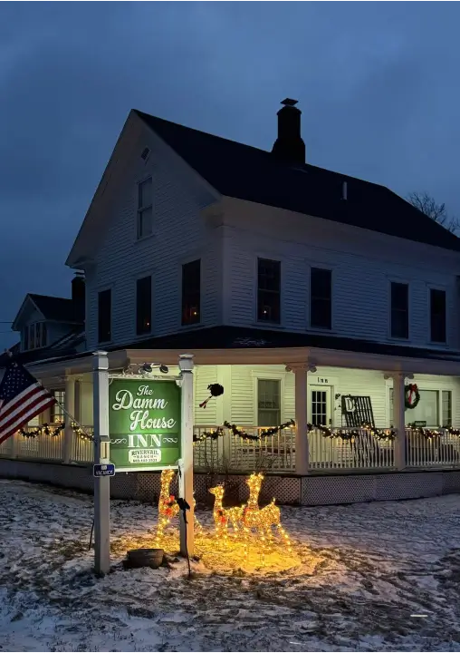 The Damn House Inn illuminated at night with festive lights, surrounded by winter scenery and American flag, highlighting a cozy retreat atmosphere.