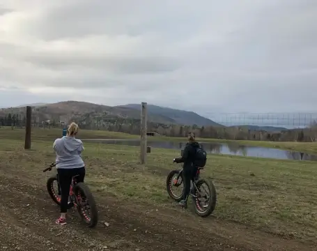 Two women biking along a dirt trail at RiverVail Ranch, with scenic hills and a pond in the background, representing outdoor recreational activities available for visitors.